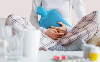 image illustrant une femme assise sur un canapé avec une bouillotte sur le ventre et des médicaments anti douleurs sur la table devant elle pour décrire l'article, gérer la douleur au quotidien avec l'endométriose