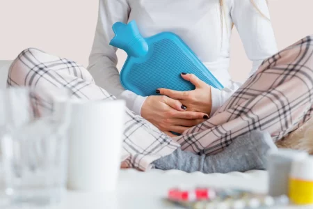 image illustrant une femme assise sur un canapé avec une bouillotte sur le ventre et des médicaments anti douleurs sur la table devant elle pour décrire l'article, gérer la douleur au quotidien avec l'endométriose